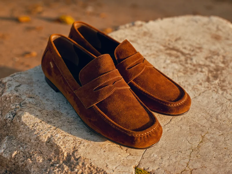 Brune ruskindsloafers med synlige syninger på en lys stenplade